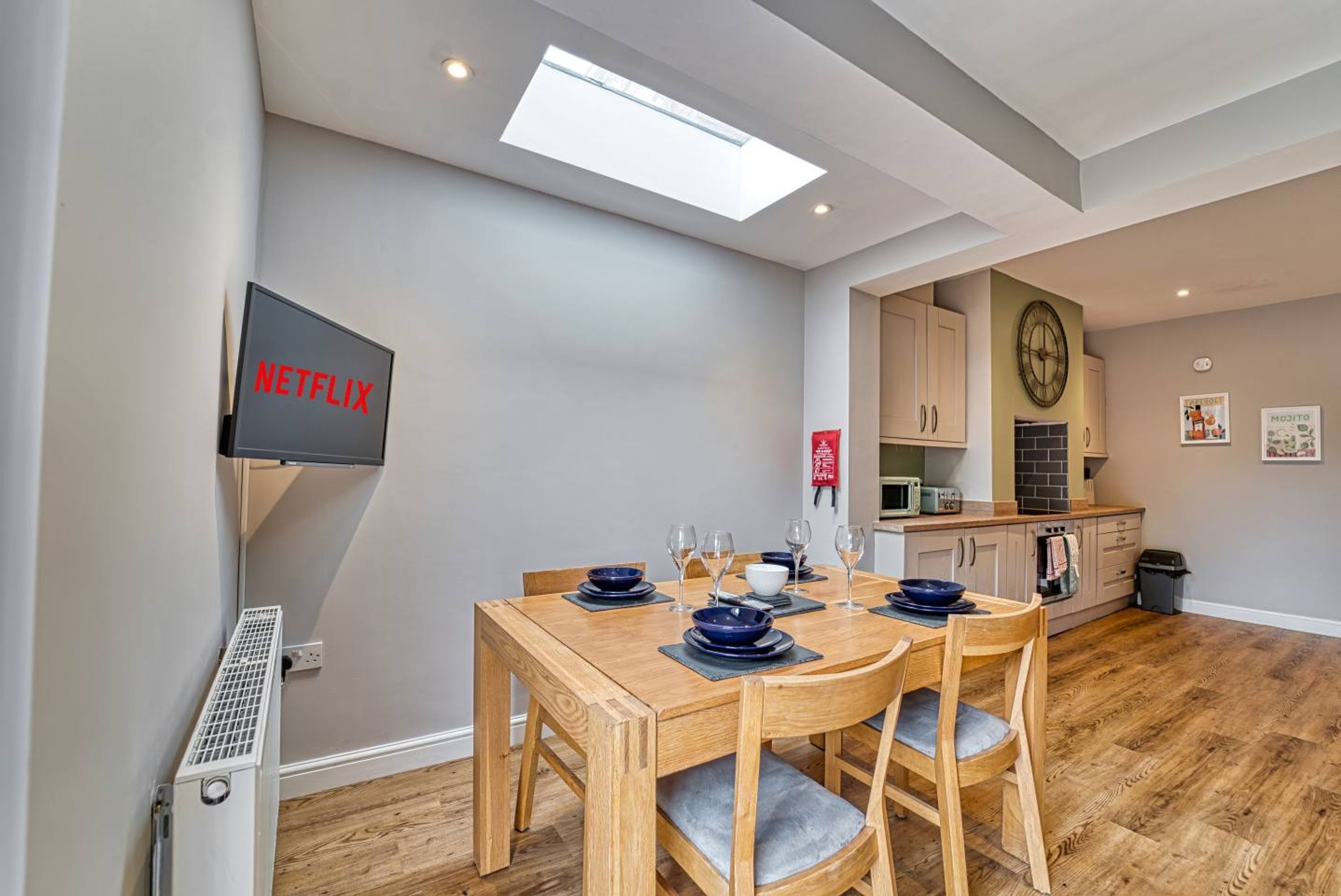 A dining area with a wooden table and chairs, a TV mounted on the wall, and a kitchen in the background.