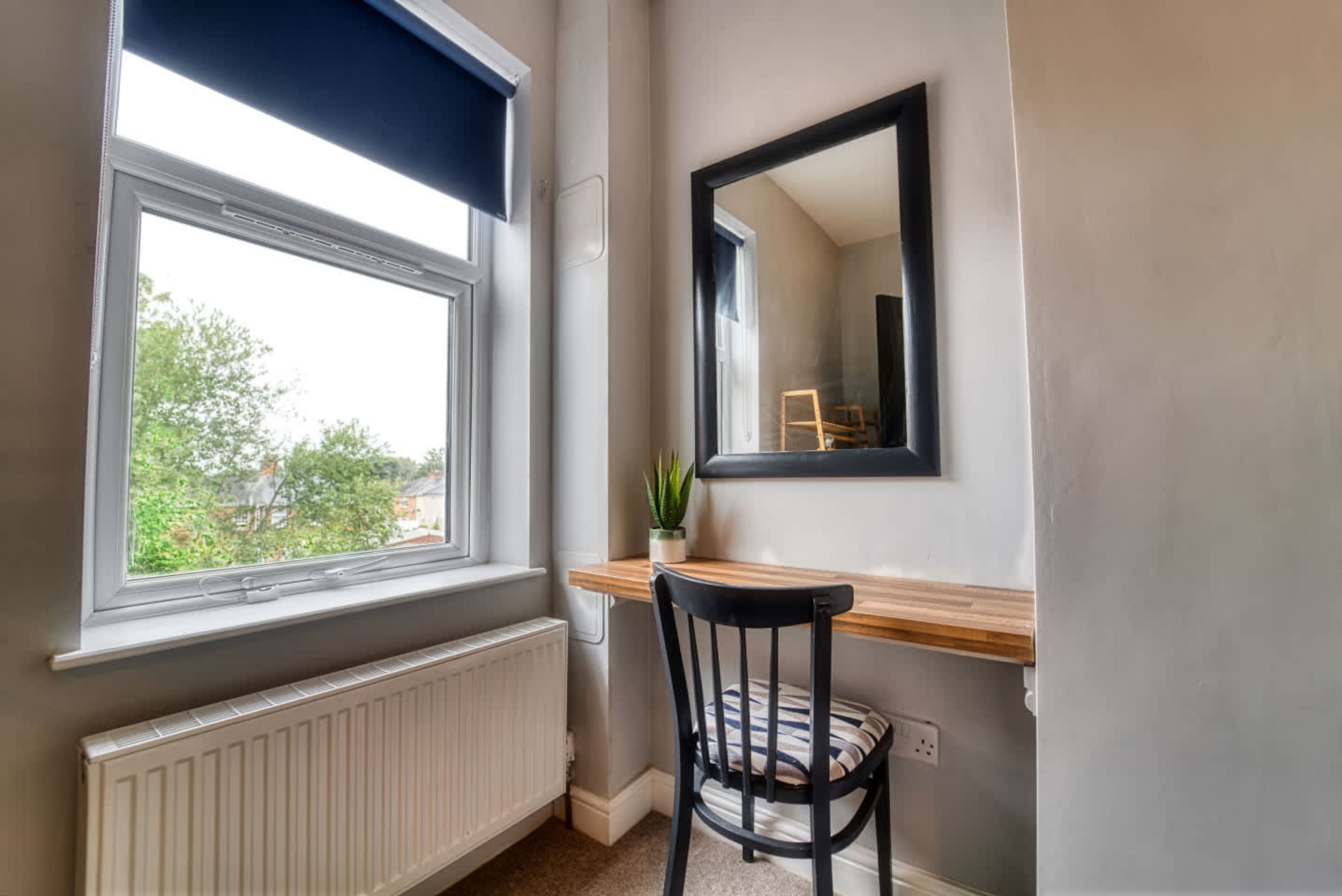 A small, well-lit room with a window, radiator, wooden desk, chair, and a mirror on the wall.