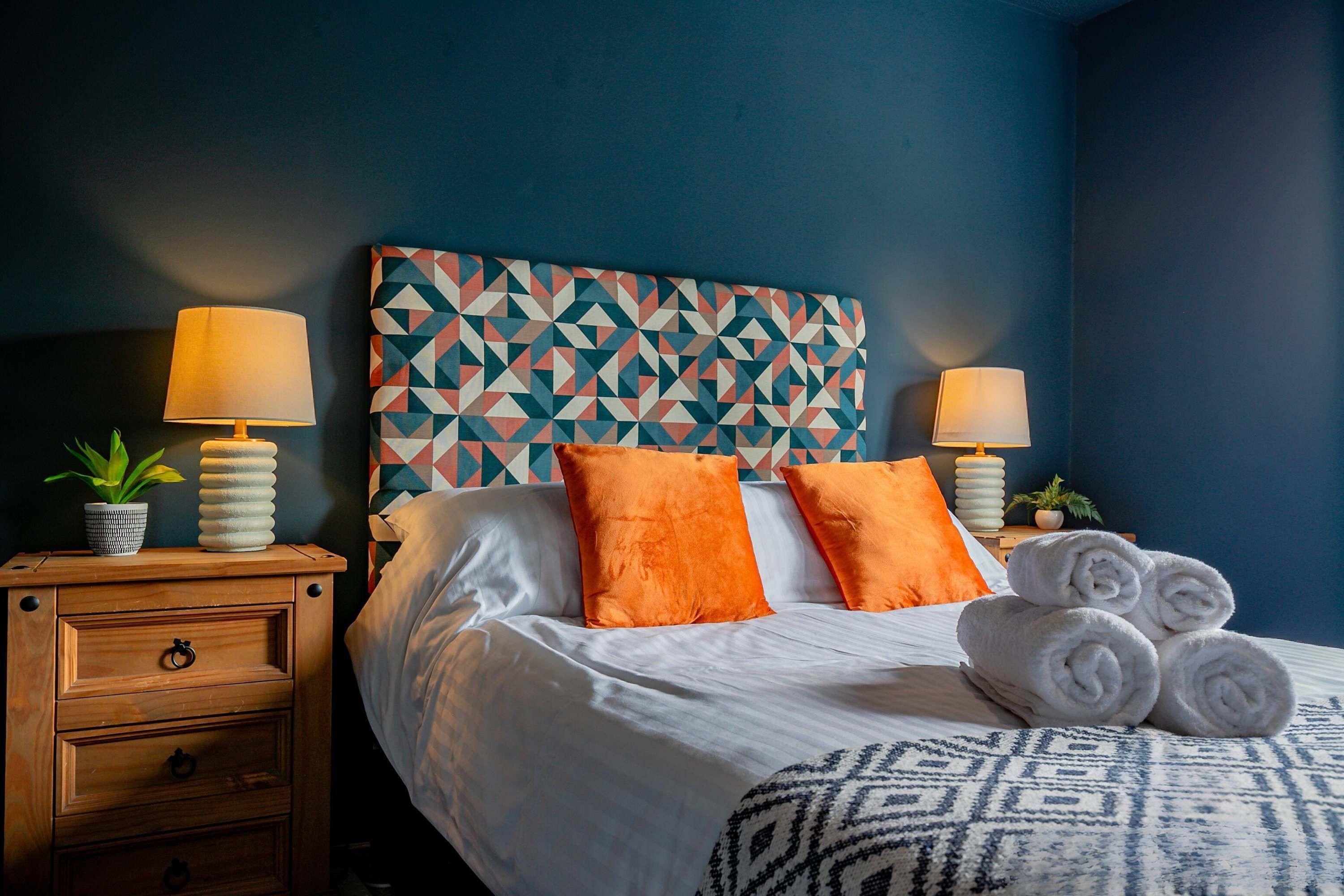 A neatly made bed with white linens, two orange pillows, and rolled towels on a patterned bedspread. There is a wooden nightstand with a lamp and a small potted plant.