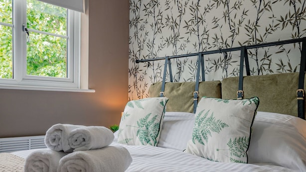Cama bem arrumada com toalhas dobradas, janela com vista para o verde e cabeceira com almofadas estampadas com folhas.