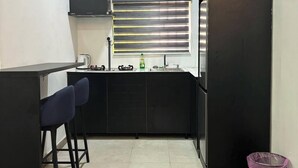 A compact kitchen with a black countertop, two bar stools, a window with blinds, a sink, and a trash bin.