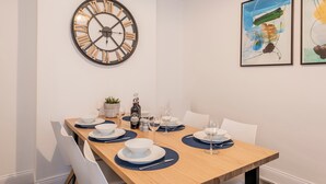 A dining area with a wooden table set for four, white chairs, a large wall clock, and two abstract paintings. - Host Stay The Haven at Church Road (Great Yarmouth)