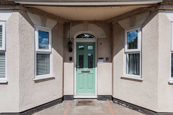 A building entrance with a green door, a welcome mat, and two windows with shutters. - Host Stay The Haven at Church Road (Great Yarmouth)