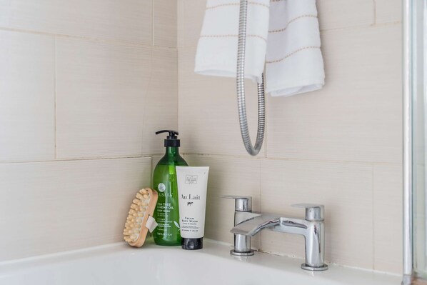 Une salle de bain avec une baignoire, une bouteille verte de gel douche, un tube de gel douche, une brosse en bois et une serviette suspendue à un porte-serviettes.