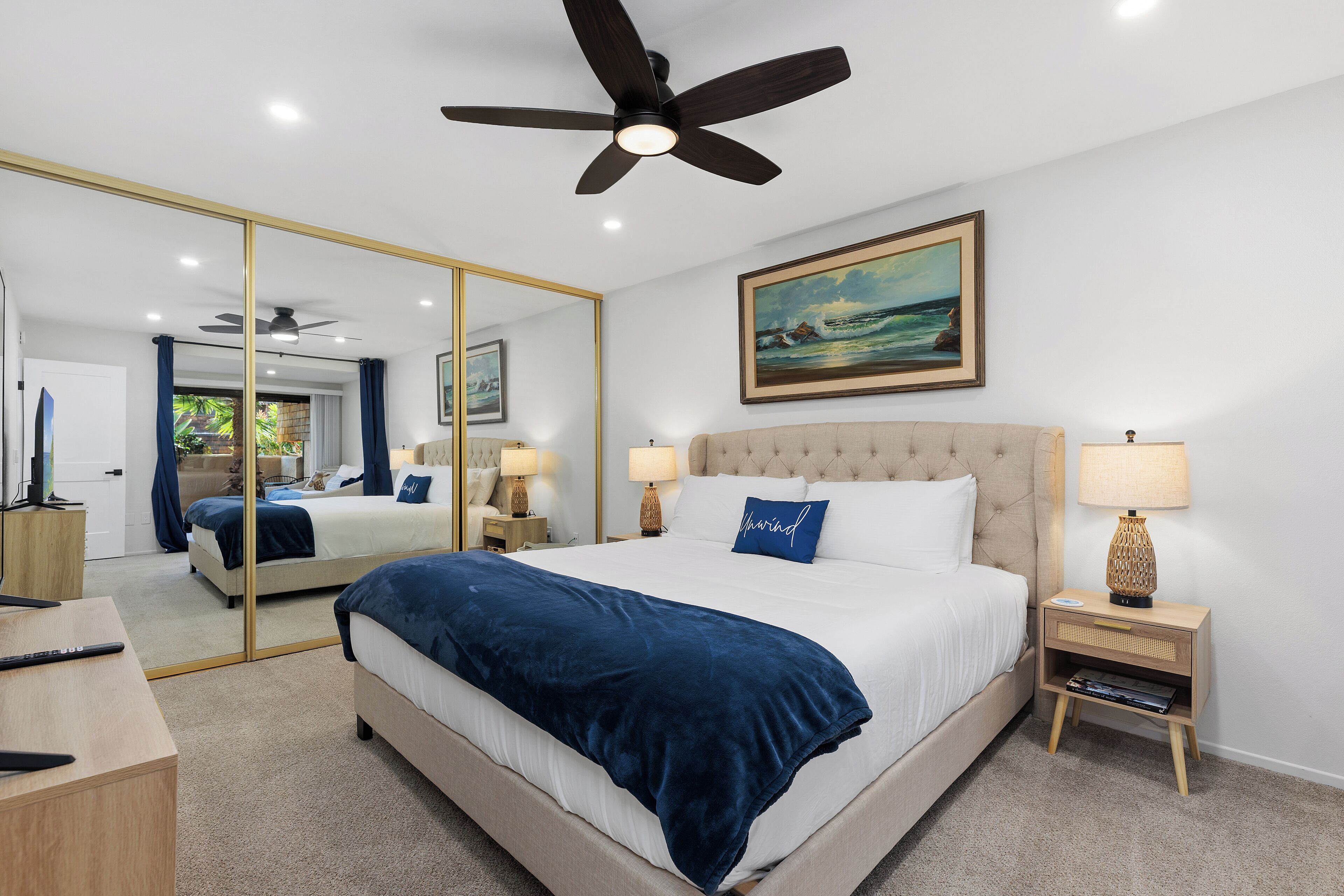A hotel room with a large bed, a ceiling fan, a painting, a bedside table with a lamp, and a mirror.