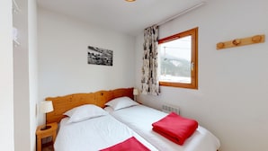 Une chambre avec un lit en bois, du linge de lit blanc, une serviette rouge, une table de chevet avec une lampe, une fenêtre avec des rideaux et un tableau accroché au mur.