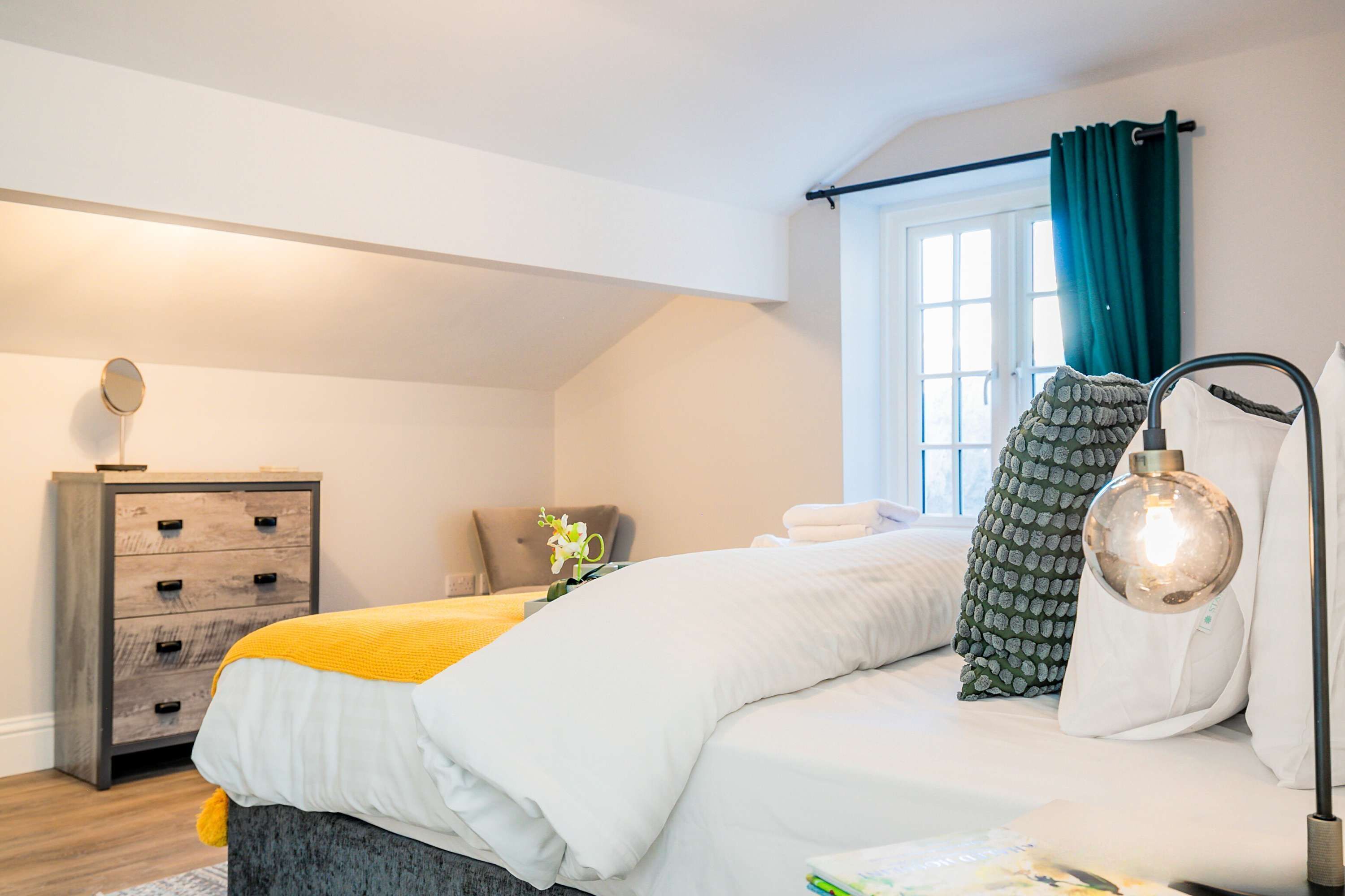 Ein Schlafzimmer mit Bett, Kommode, einem Fenster mit Vorhängen und einer Lampe.