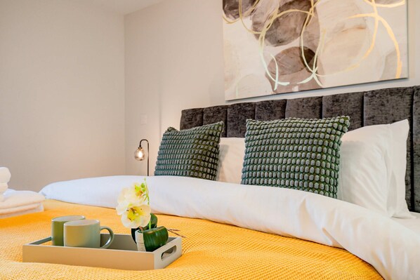 A neatly made bed with a tray of mugs and a flower, a modern headboard, and a wall art piece.