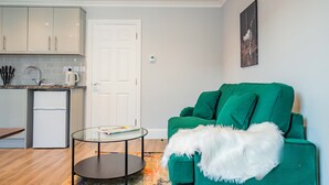 Ein kompakter Wohnbereich mit Küchenzeile, einem grünen Sofa, einem Glastisch und einer weißen Tür.