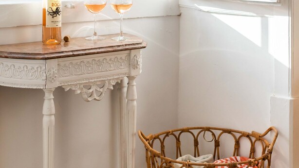 Une pièce avec une table blanche près d’une fenêtre, un panier en osier rempli de serviettes et une bouteille de vin.