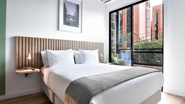 A modern bedroom with a large bed, a wooden headboard, a bedside table with a lamp, a framed picture on the wall, and a window with a view of buildings. - w Radiant Loft w Balcony in Chapinero (Bogotá)