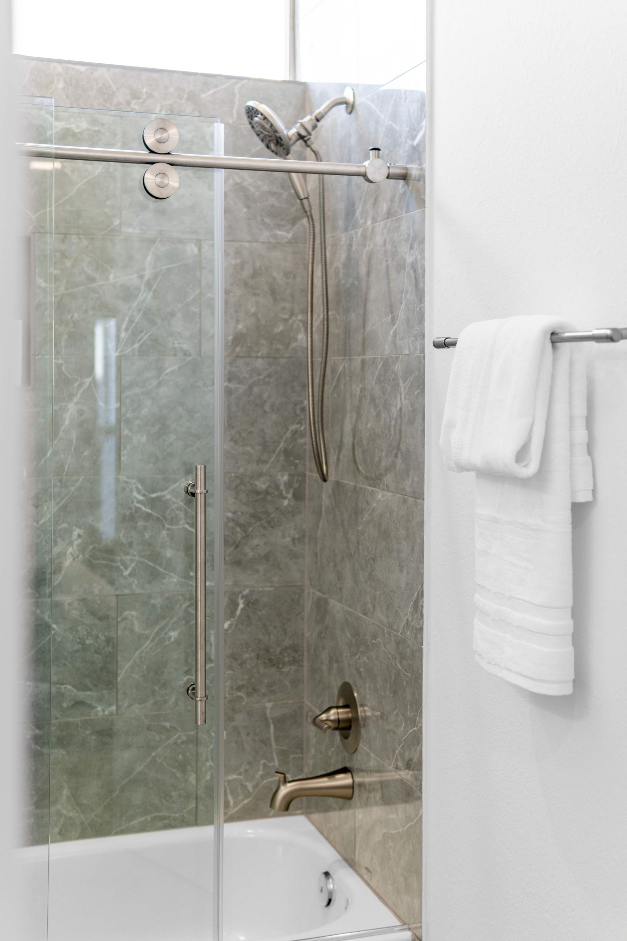 A glass-enclosed shower stall with a handheld showerhead, a wall-mounted towel rack with a towel, and a bathtub with a faucet.