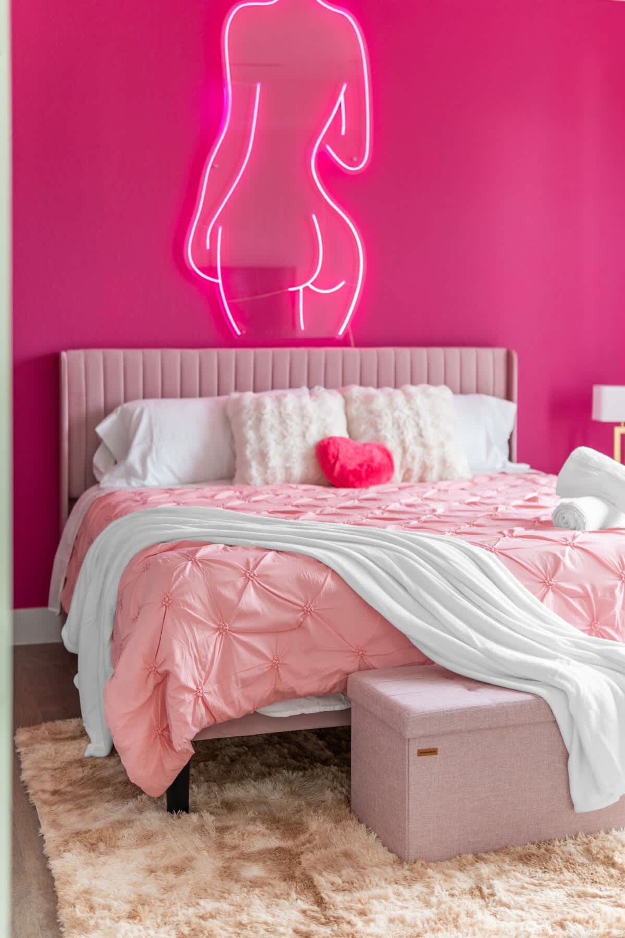 A bedroom with a pink bedspread, a fluffy rug, and a neon silhouette on the wall.