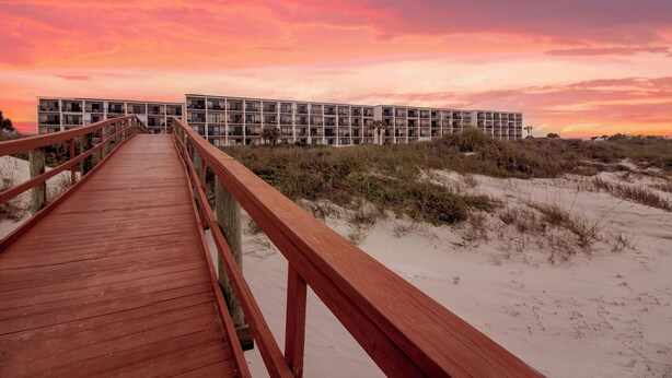 Une promenade en bois mène à un bâtiment moderne doté de nombreux balcons, situé sur une plage de sable avec des dunes en arrière-plan.
