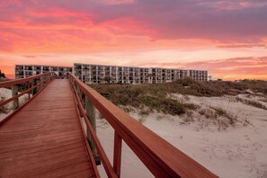A wooden boardwalk leading to a modern building with multiple balconies, situated on a sandy beach with dunes in the background. - Beacher's Oceanfront Patio, Condo Complex Construction 10/2026-1/2027 (St. Augustine)
