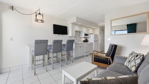 Un salon moderne avec un canapé gris, une table basse blanche et un coin repas avec des chaises grises. On aperçoit à l’arrière-plan une cuisine avec des meubles blancs et un téléviseur à écran plat.