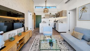 Una cocina moderna con una isla central, un comedor con una mesa de centro de vidrio y una sala de estar con un sofá y televisor.