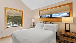 A bedroom with a large bed, a nightstand with a lamp, a window with blinds, and a mirror above the bed.
