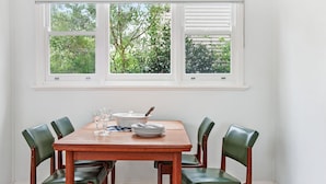 A dining area with a wooden table and four chairs, a window with three panes, and a view of greenery outside.