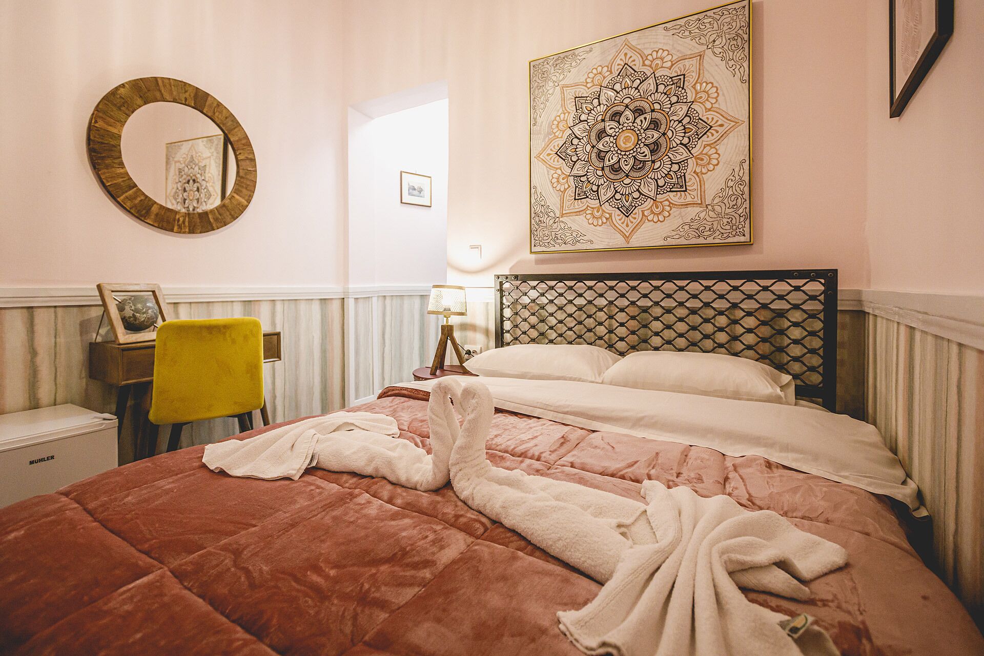 A hotel room with a bed, a round mirror, a framed picture, a yellow chair, and a decorative wall piece.