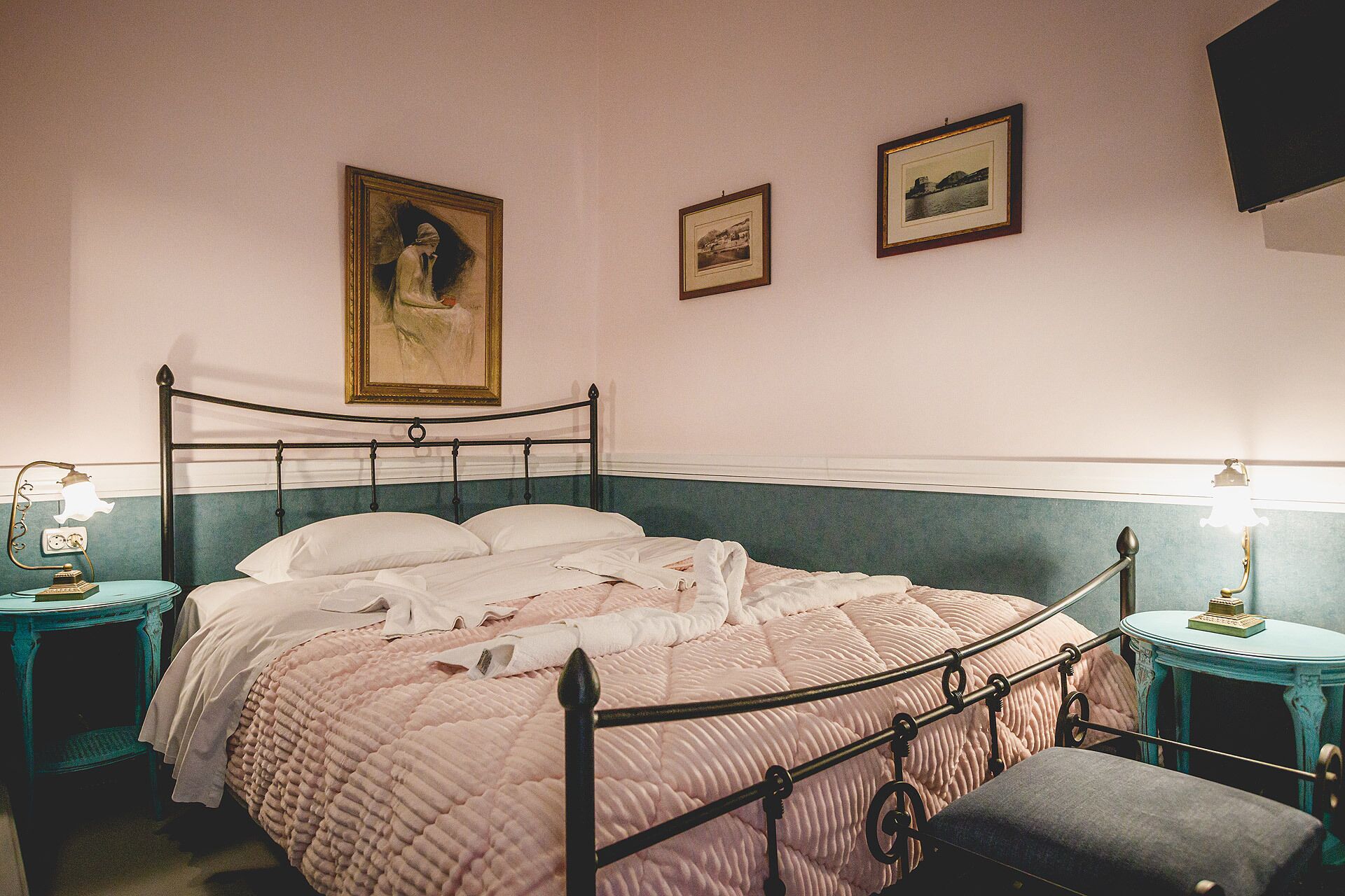 A neatly made bed with a pink quilt, two bedside tables with lamps, and three framed pictures on the wall.