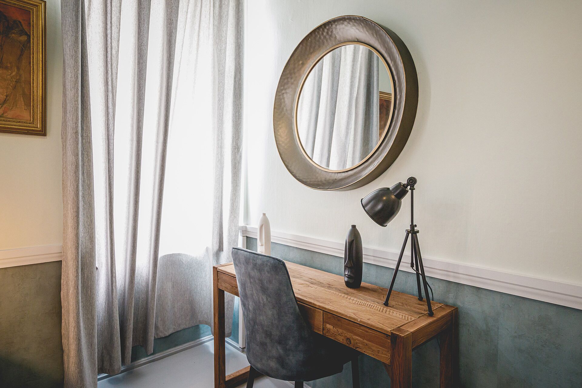 A room with a wooden desk, a black chair, a round mirror, and a desk lamp on a tripod.