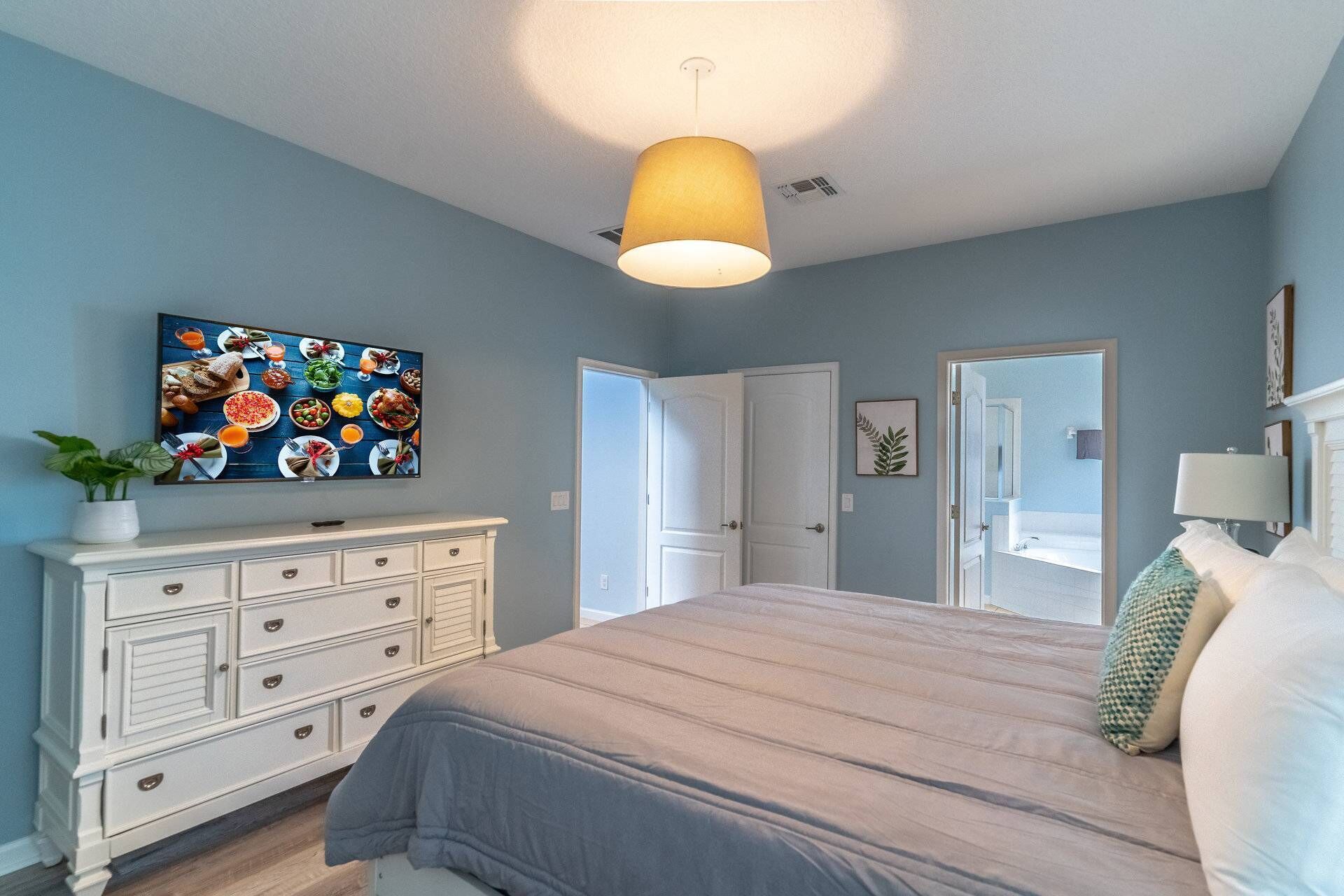 A bedroom with a bed, a dresser, a TV on the wall, and a door leading to a bathroom.