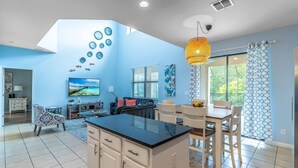 A modern kitchen with a central island, a dining area with chairs, a TV in the living room, and a large window with curtains. - 2614 Veranda Palms (Kissimmee)