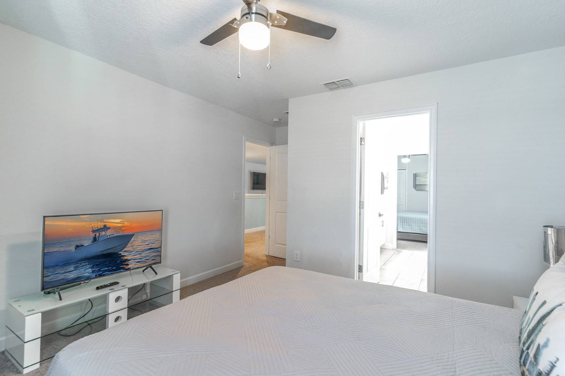 A bedroom with a bed, a TV on a stand, a ceiling fan, and a door leading to another room.