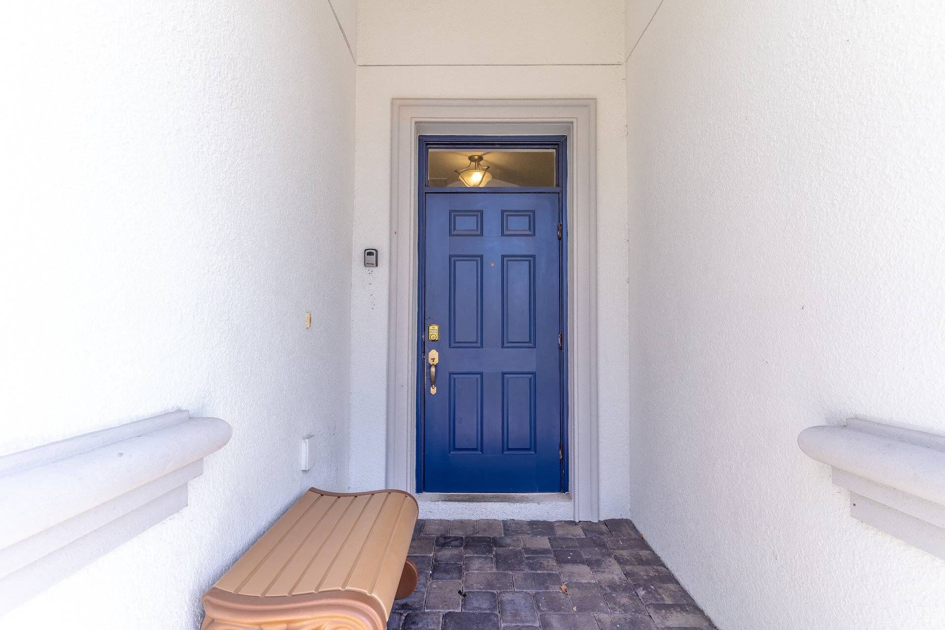 A blue door with a light fixture above it, a bench in front, and a white wall with a small window to the left.