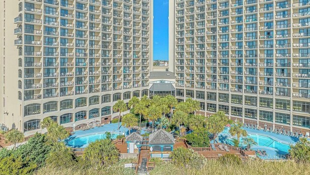 A high-rise hotel with multiple balconies, a central pool area, and surrounding greenery. - 🌊Oceanfront | Amazing Views | Fabulous Amenities | Beach Cove Resort 1020⛱️ (North Myrtle Beach)