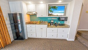 A compact kitchen with white cabinets, a stainless steel refrigerator, a granite countertop, and a built-in TV displaying a beach scene. - 🌊Oceanfront | Amazing Views | Fabulous Amenities | Beach Cove Resort 1020⛱️ (North Myrtle Beach)