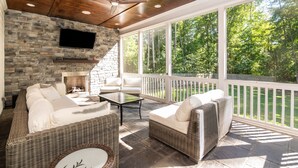 A covered patio with wicker furniture, a stone wall, a flat-screen TV, and a view of the forest. - Durham Retreat Games, Exercise, And Relaxation 5 Bedroom Home (Durham)