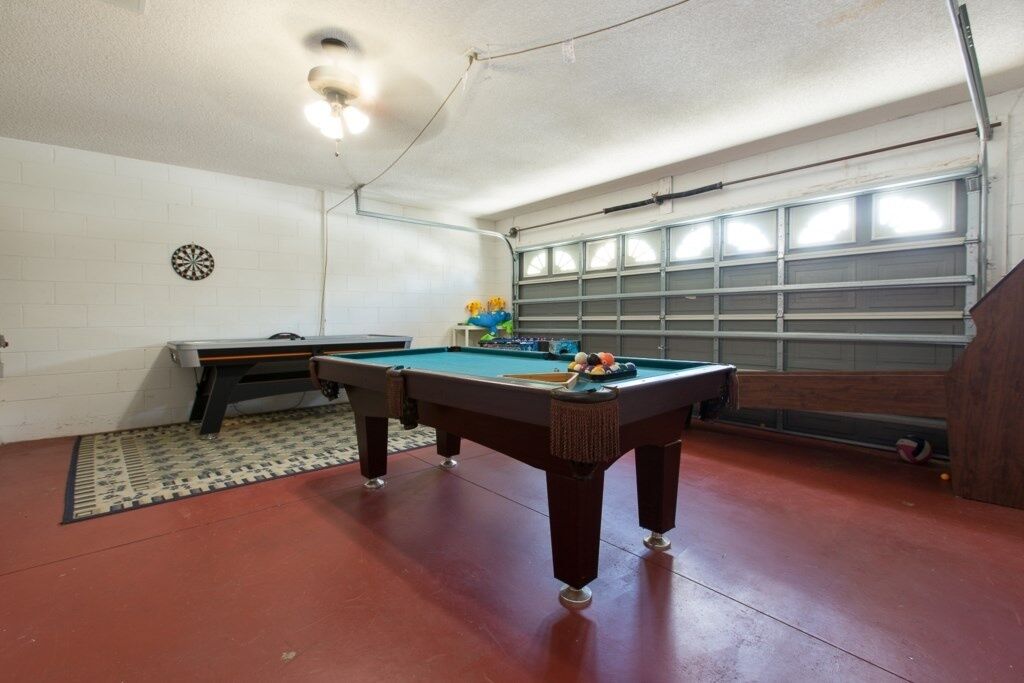 Una mesa de billar en una habitación con piso rojo, un dardo y una puerta de garaje al fondo.