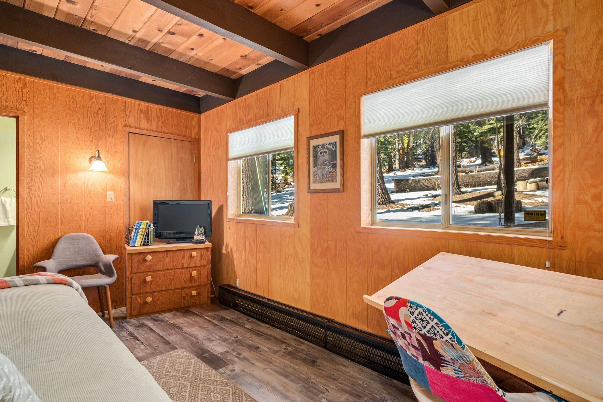 Acogedora habitación tipo cabaña con paredes de madera, televisión, una silla, cómoda y ventanas grandes con vistas a un paisaje nevado.