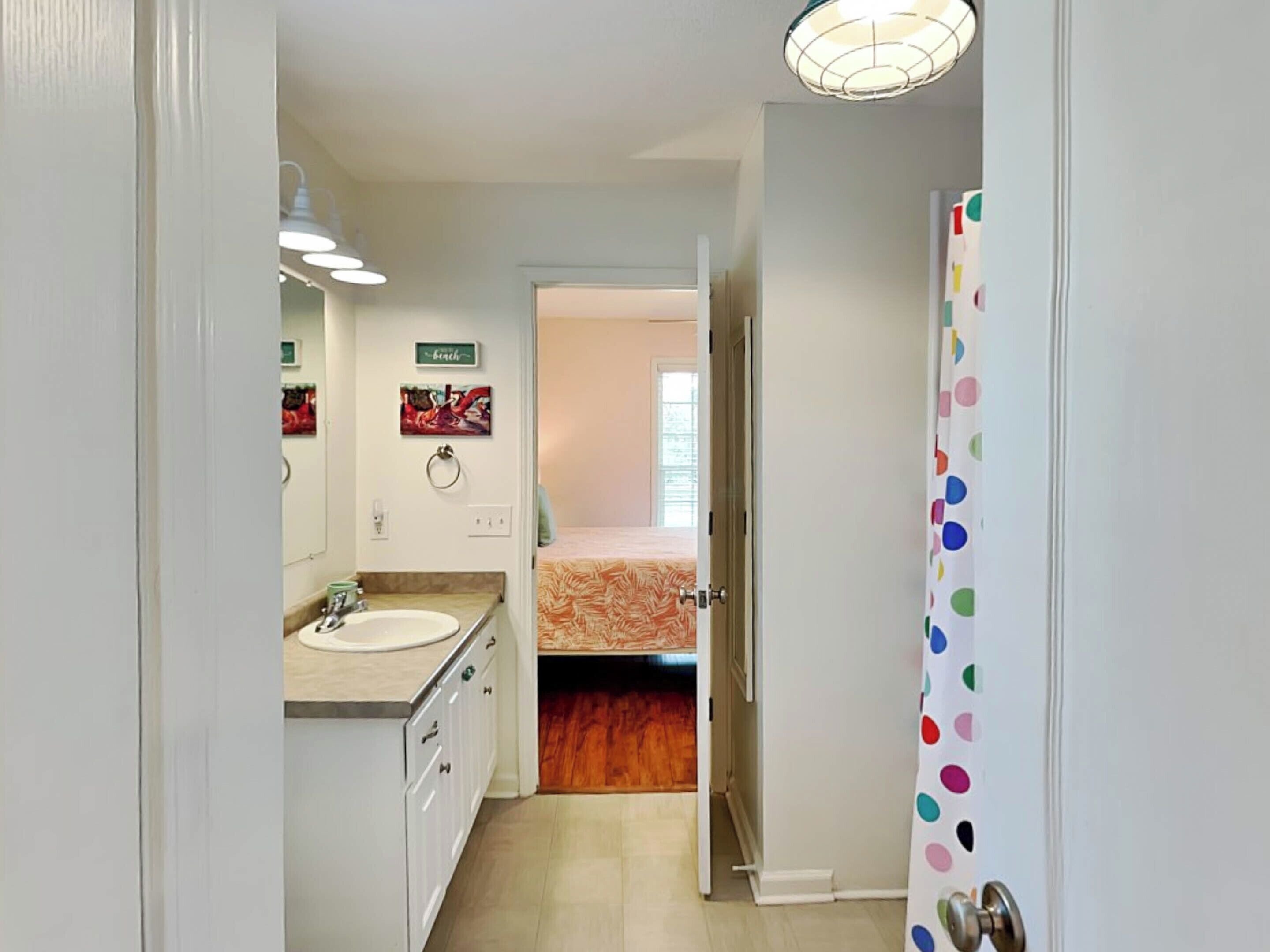 A bathroom with a sink, mirror, and towel rack. There is a door leading to a bedroom with a bed and a window.