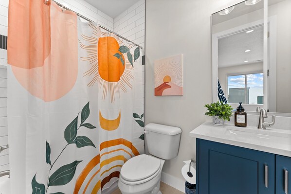 Une salle de bain moderne avec des toilettes blanches, un meuble bleu et un rideau de douche orné de motifs représentant un soleil et des feuilles.
