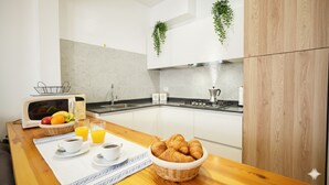 Eine moderne Küche mit weißen Schränken, einer Mikrowelle, einem Spülbecken und einem Herd. An der Wand stehen zwei Blumentöpfe, ein Korb mit Croissants und ein Tisch mit Untersetzer, zwei Kaffeetassen und einem Glas Orangensaft.