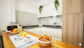 A modern kitchen with white cabinets, a microwave, a sink, and a stove. There are two potted plants on the wall, a basket of croissants, and a table with a placemat, two cups of coffee, and a glass of orange juice. - Appartamento Arcobaleno (Costarainera)