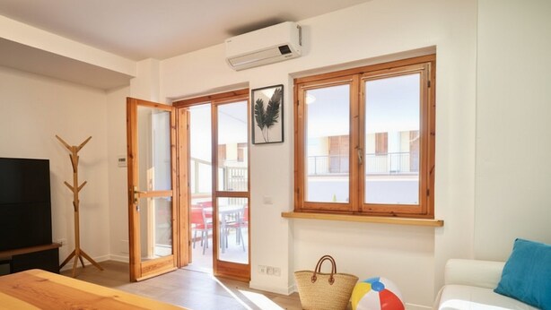 Ein Wohnzimmer mit Couch, blauem Kissen, Couchtisch aus Holz, Fernseher und einem Strandball. Zwei Fenster mit Holzrahmen und eine Tür zu einem Außenbereich mit Tisch und Stühlen.