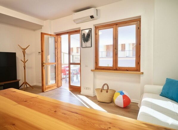 Una sala de estar con una mesa de centro de madera, un televisor, un sofá con una almohada azul y una pelota de playa. Hay dos ventanas con marcos de madera y una puerta que da a un área exterior con una mesa y sillas.