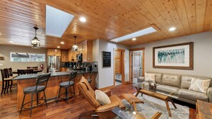 A spacious living area with a wooden ceiling, a bar with stools, a sofa, and a large painting on the wall.