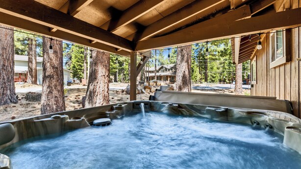 A hot tub area with a wooden deck, surrounded by trees and a view of a cabin.