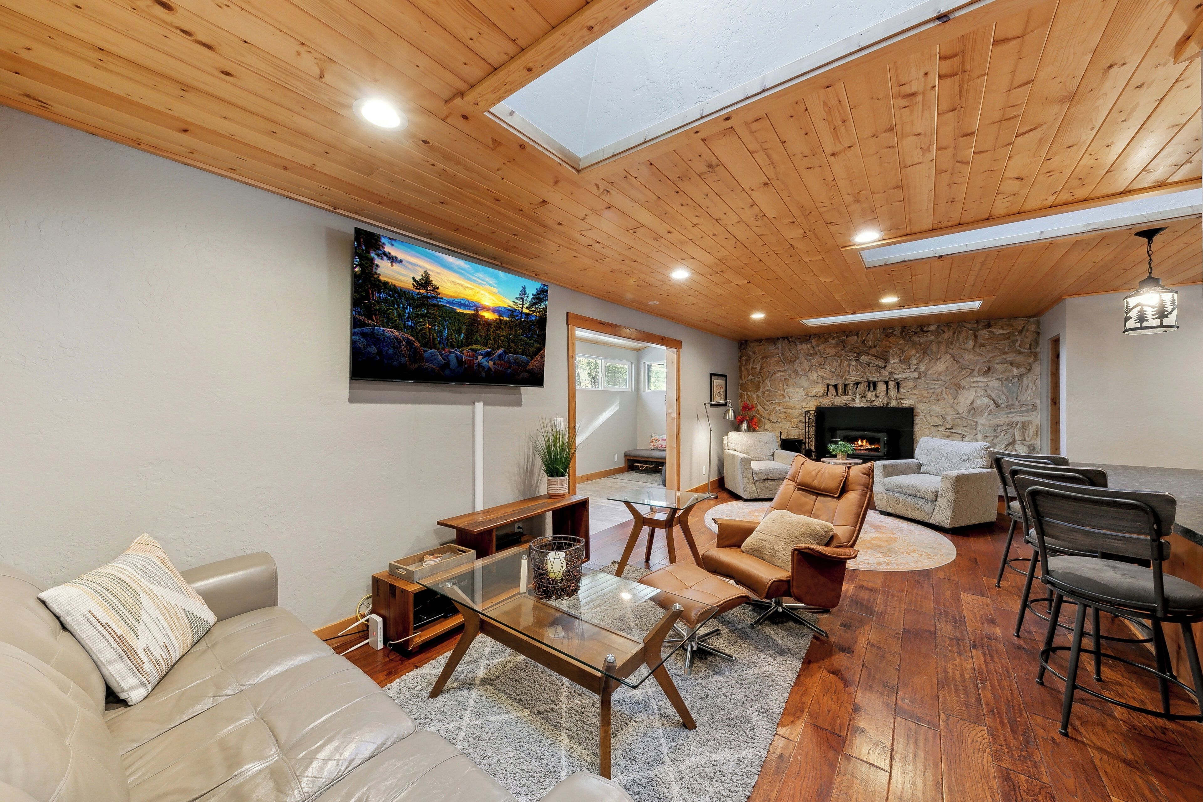 A modern living room with a stone fireplace, wooden ceiling, and a large TV mounted on the wall.