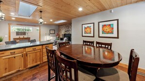 A wooden dining area with a large table and chairs, a kitchen with wooden cabinets, a stone wall, and two framed paintings on the wall.