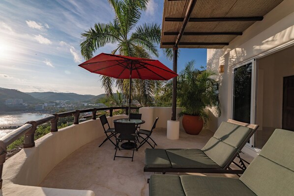 A balcony with a table and chairs, a red umbrella, and a view of the sea and mountains.