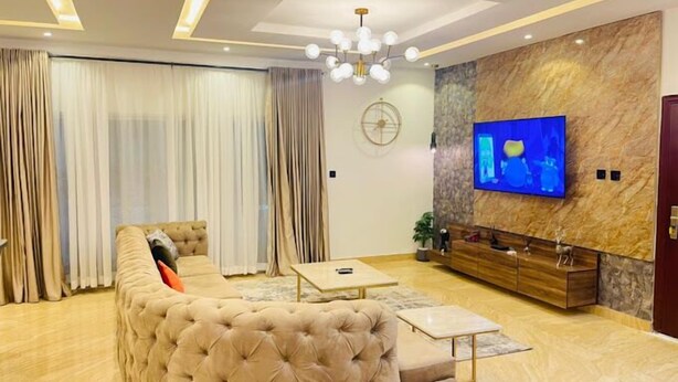 Uma sala de estar moderna com um amplo sofá modular, uma mesa de centro, uma TV fixada numa parede de mármore e um candeeiro de lustre.