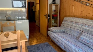 Un espace de vie compact comprenant un coin cuisine, un canapé recouvert d’un tissu à motifs et une porte en bois donnant sur une autre pièce.