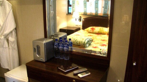 A compact hotel room with a small desk, a microwave, a scale, a remote control, and a mirror reflecting a bed with a floral bedspread. - LIS CHAIN HOSTEL (Kowloon)