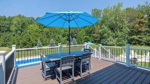 Una terraza de madera con una mesa, sillas, un paraguas azul y una piscina al fondo.